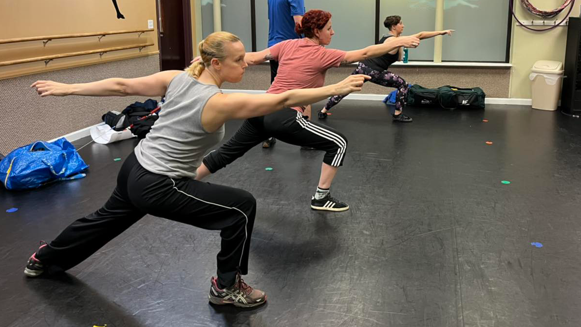 Group rapier class. Several women rapier fencers working on their lunges, practicing in front of a mirror.